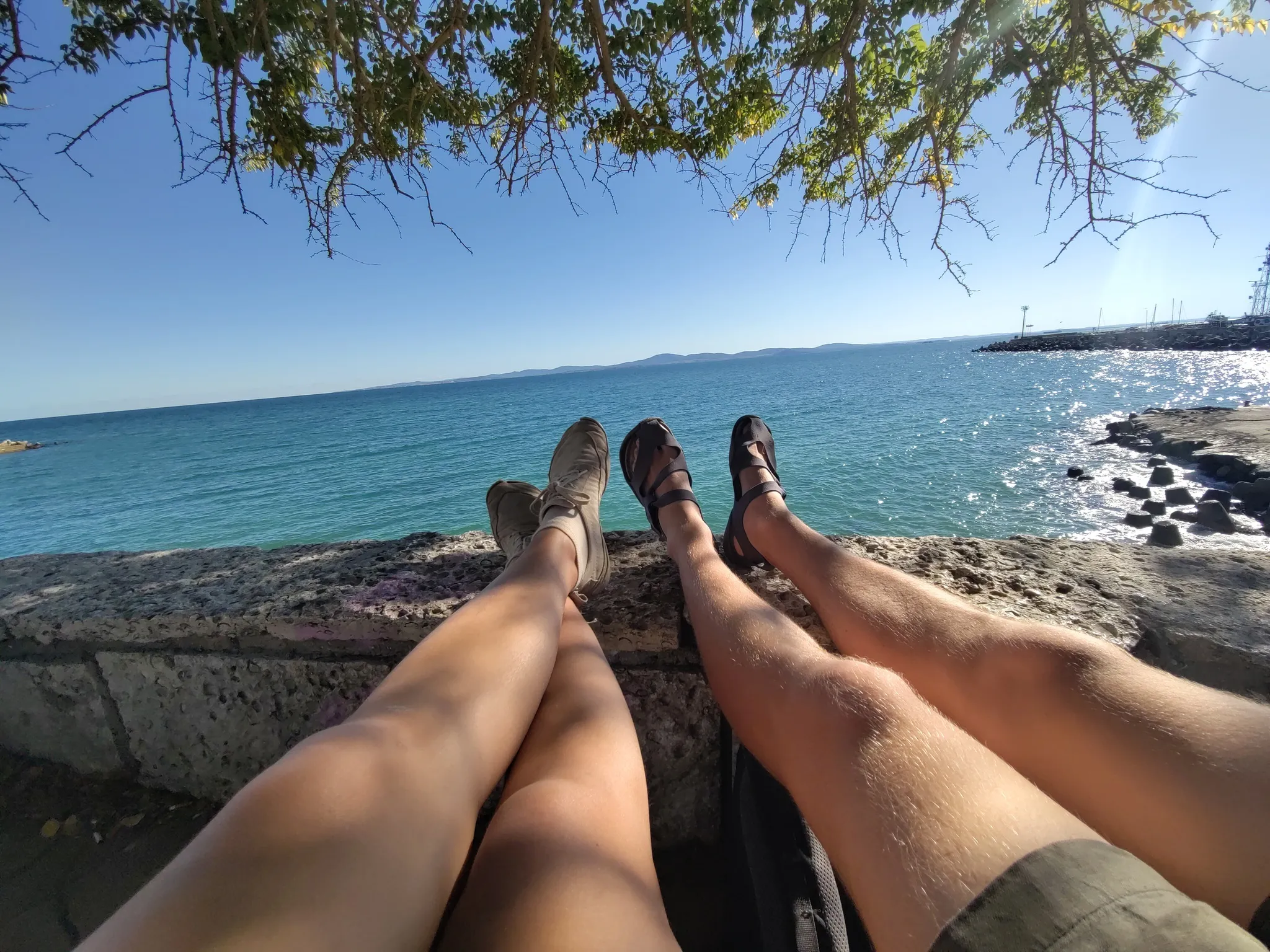 Chiara und Tim haben ihre Beine auf einem Steinwall liegen. Im Hintergrund sieht man das Meer.