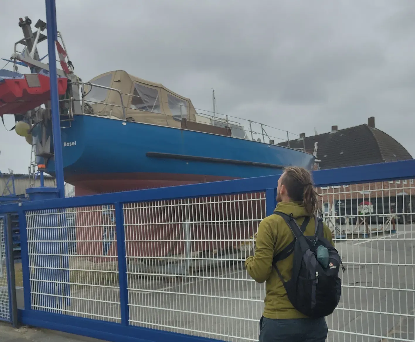 Tim steht vor dem Tor der Werft und guckt auf das direkt auf der andere Seite befindliche Boot.