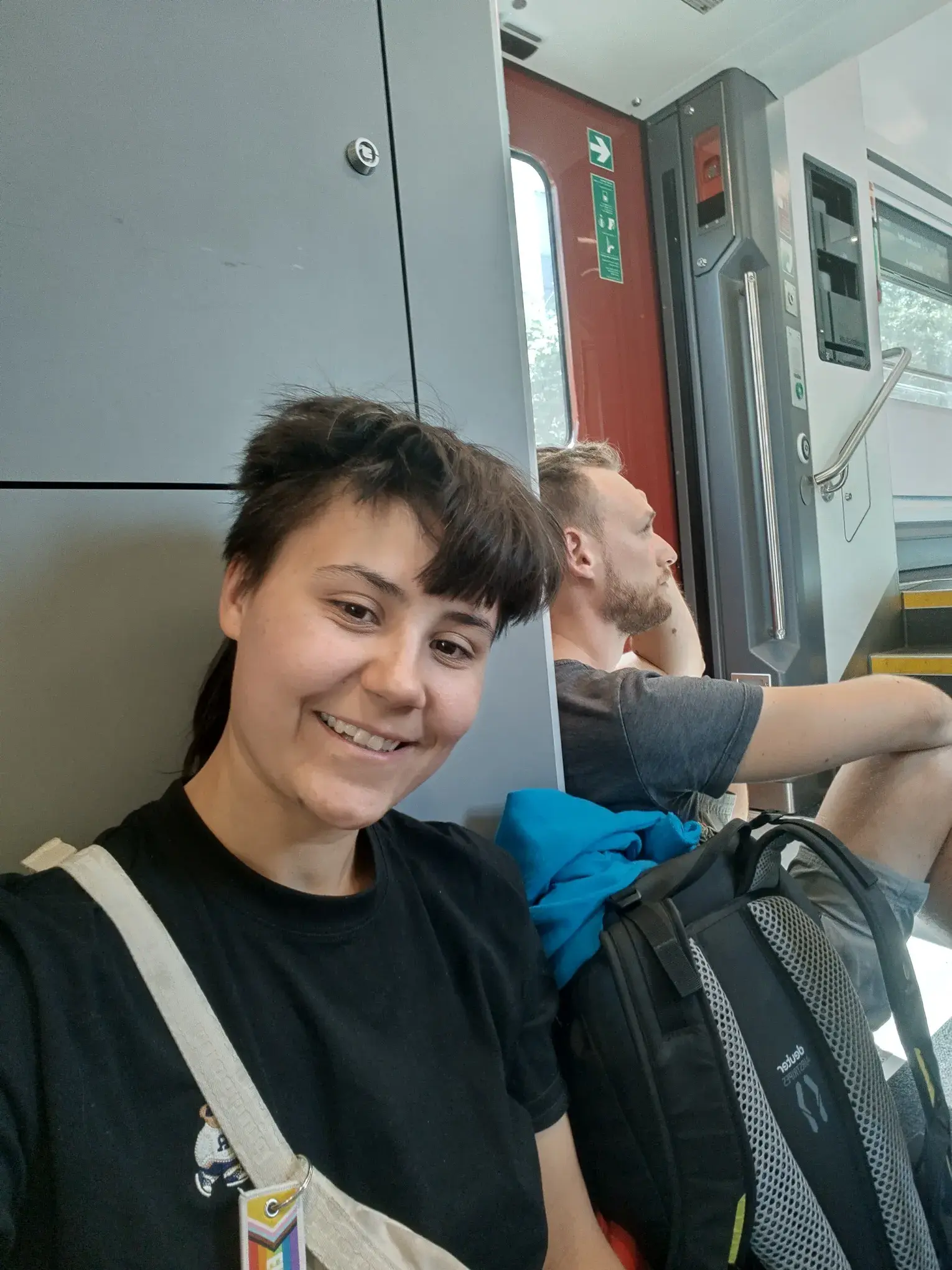 Chiara macht ein Selfie auf dem Boden eines RailJet sitzend. Tim guckt im Hintergrund grimmig.