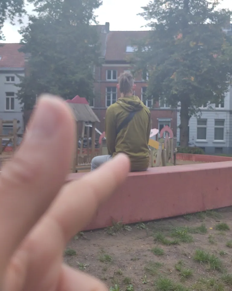 Chiara zielt mit entsichertem Finger auf Tim, der auf einer Bank sitzt und den Spielplatz anguckt.